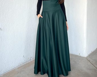 May include: A long, dark green skirt with a high waist and pockets, paired with a black long-sleeved top. The skirt has a flowing, full shape. The outfit is set against a white wall.