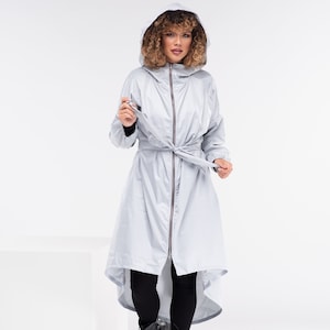 Puede incluir: Un impermeable con capucha de color gris claro con cremallera y cinturón anudado. El abrigo tiene un dobladillo alto-bajo y se usa con leggings negros y botas negras con cordones. La modelo lleva una camiseta negra de manga larga.