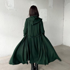 May include: A long, dark green hooded coat with a belt. The coat is flowing in the wind and the hood is up.