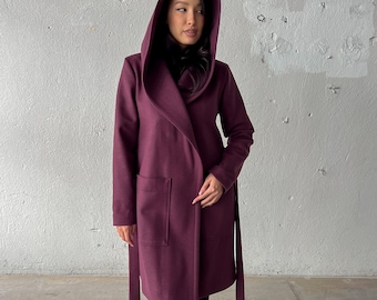 May include: A burgundy hooded coat with a wrap-style closure and a matching belt. The coat features two front pockets and is worn with black leggings and black combat boots. The background is a textured, light gray wall.