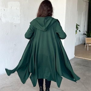 Può includere: Un cardigan con cappuccio verde scuro in stile mantello, con maniche lunghe e orlo asimmetrico fluente. Il cardigan è realizzato in un tessuto morbido e viene indossato con pantaloni e stivali neri. La persona è di spalle.