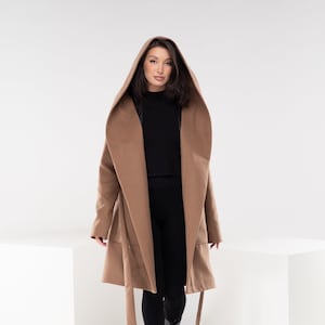 May include: A brown hooded coat with a tie belt. The coat is worn by a person with long dark hair and is standing on a white surface.