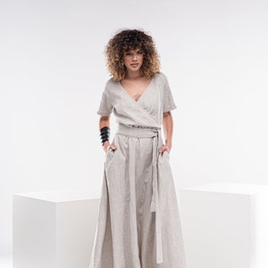 May include: A woman wearing a light beige linen wrap dress with a long skirt and short sleeves. She is standing on a white platform with her hands in her pockets.