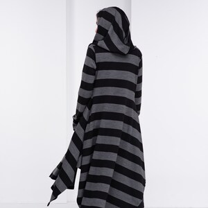 Op de afbeelding: Een lange jas met capuchon met horizontale zwarte en grijze strepen. De jas heeft een vloeiende, asymmetrische zoom en wordt gedragen met een zwarte broek en laarzen. De capuchon is omhoog en de rug van de persoon is naar de camera gericht.