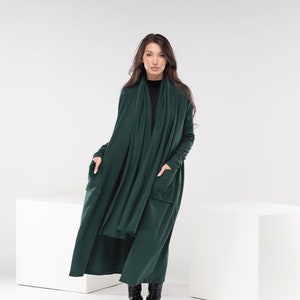 Könnte beinhalten: Ein langer, dunkelgrüner Cardigan mit langen Ärmeln und Taschen. Der Cardigan wird über einem schwarzen Rollkragenpullover getragen und das Model trägt schwarze Stiefel.
