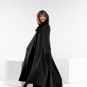 May include: A black hooded cape with a fur trim around the hood. The cape is flowing in the wind and the model is looking over her shoulder.
