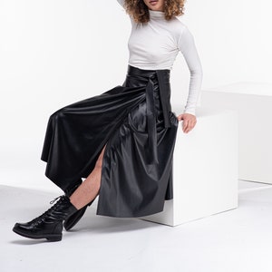 May include: A woman wearing a white turtleneck shirt and a black leather wrap skirt. She is sitting on a white cube and wearing black combat boots.