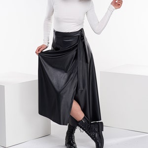 May include: A woman wearing a white turtleneck top and a black faux leather wrap skirt. She is wearing black lace-up boots and has curly brown hair.