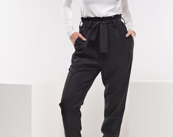May include: A woman wearing a white turtleneck shirt and black high-waisted pants with a tie belt. She is wearing black ankle boots.