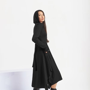 Könnte beinhalten: Ein schwarzes Kapuzenkleid mit langem Rock und Taillengürtel. Das Kleid hat einen langen, fließenden Rock mit einem leicht asymmetrischen Saum. Das Model trägt schwarze Schnürstiefeletten.