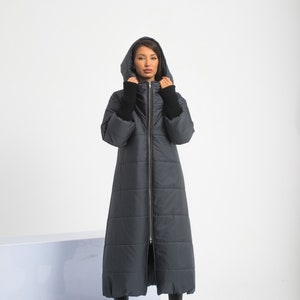 Puede incluir: Una larga chaqueta acolchada de color gris oscuro con cierre de cremallera y capucha. La chaqueta tiene un diseño acolchado y la lleva puesta una persona con botas negras.