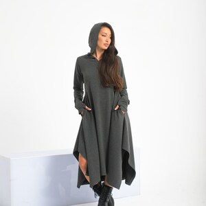 Könnte beinhalten: Ein langes, graues Kapuzenkleid mit hohem Ausschnitt und langen Ärmeln. Das Kleid hat einen fließenden, asymmetrischen Saum und Taschen an den Seiten. Das Model trägt schwarze Stiefel.