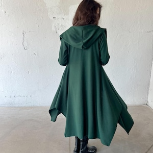May include: A long, flowing, forest green hooded coat. The coat features an asymmetrical hemline and long sleeves. The person is wearing black boots. The coat is made of a soft, draping fabric.