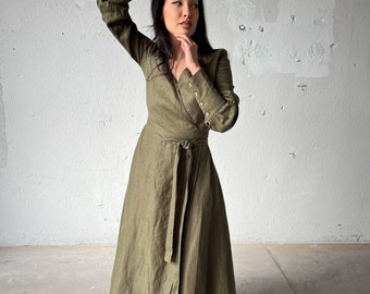 Puede incluir: Una mujer lleva un vestido largo de color verde oliva con un cinturón de anudar. El vestido tiene mangas largas con detalles de botones en los puños. Está de pie frente a una pared blanca.