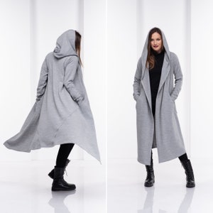 Op de afbeelding: Een lange, grijze hooded cardigan met een gedrapeerd ontwerp. De cardigan heeft een losse pasvorm en lange mouwen. Het model draagt de cardigan met een zwarte broek en zwarte laarzen.
