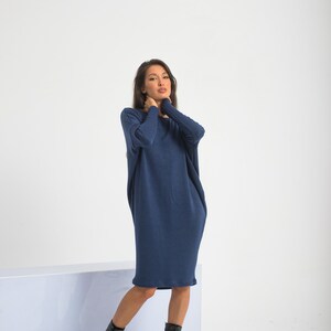 May include: A woman wearing a long, navy blue, loose-fitting dress with long sleeves. She is wearing black lace-up boots and is standing in front of a white background.