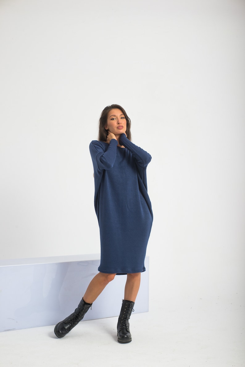 May include: A woman wearing a long, navy blue, loose-fitting dress with long sleeves. She is wearing black lace-up boots and is standing in front of a white background.