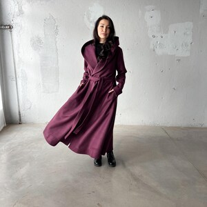 May include: A long, hooded coat in a deep burgundy color. The coat features a wrap-around design with a matching belt and pockets. The model is wearing black boots. The coat's fabric appears to be a soft, flowing material.