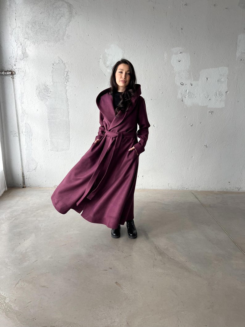 May include: A long, hooded coat in a deep burgundy color. The coat features a wrap-around design with a matching belt and pockets. The model is wearing black boots. The coat's fabric appears to be a soft, flowing material.