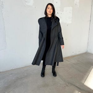 Puede incluir: Un abrigo largo de color gris oscuro con capucha y cintur&oacute;n de lazo. El abrigo est&aacute; hecho de una tela suave y c&aacute;lida y tiene un corte holgado. La modelo lleva botas negras.