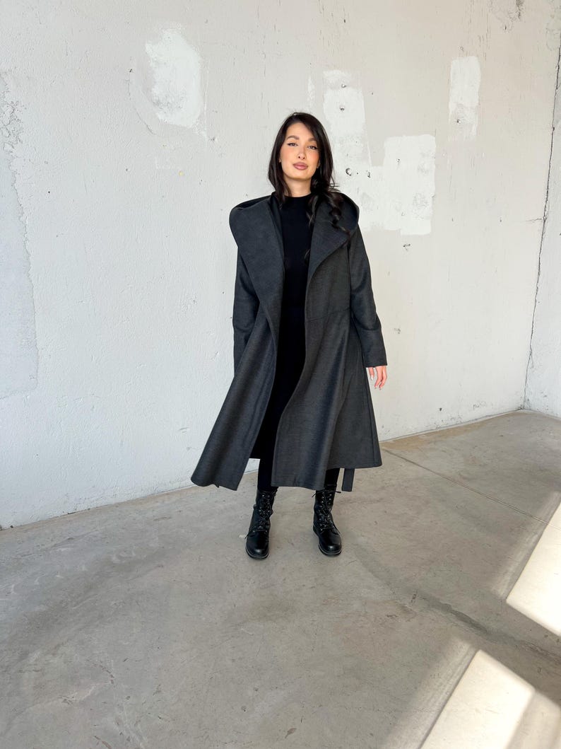Puede incluir: Un abrigo largo de color gris oscuro con capucha y cintur&oacute;n de lazo. El abrigo est&aacute; hecho de una tela suave y c&aacute;lida y tiene un corte holgado. La modelo lleva botas negras.