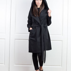 K&ouml;nnte beinhalten: Ein schwarzer Mantel mit Kapuze, Bindeg&uuml;rtel und gro&szlig;en Taschen. Der Mantel ist knielang und wird mit einer schwarzen Hose und metallisch-silbernen Schuhen getragen. Der Hintergrund zeigt wei&szlig;e T&uuml;ren und einen Holzboden.