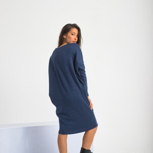 May include: A woman wearing a long, navy blue, oversized sweater dress with long sleeves. She is wearing black lace-up boots and is standing on a white surface.