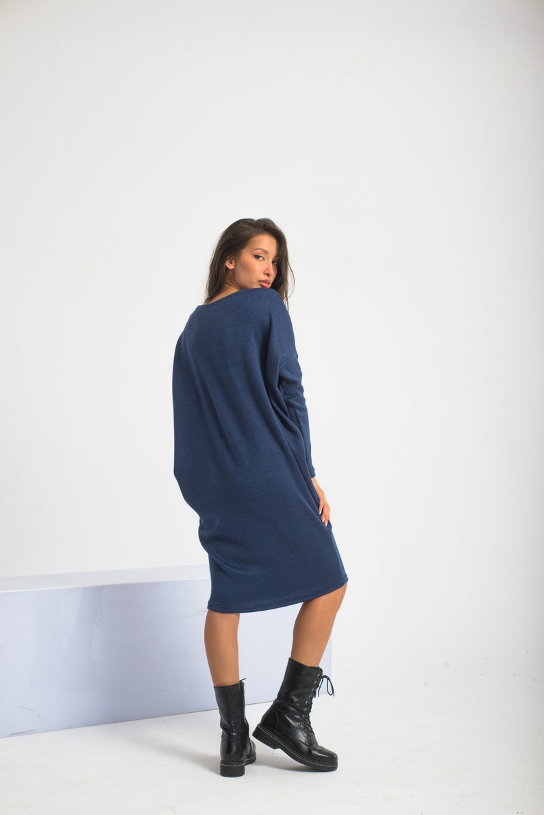 May include: A woman wearing a long, navy blue, oversized sweater dress with long sleeves. She is wearing black lace-up boots and is standing on a white surface.