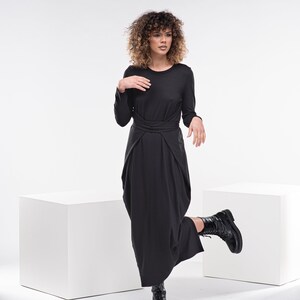 May include: A woman wearing a black long-sleeved dress with a knotted waist. She is standing on one leg with her other leg bent at the knee and her foot resting on a white cube. The background is white.