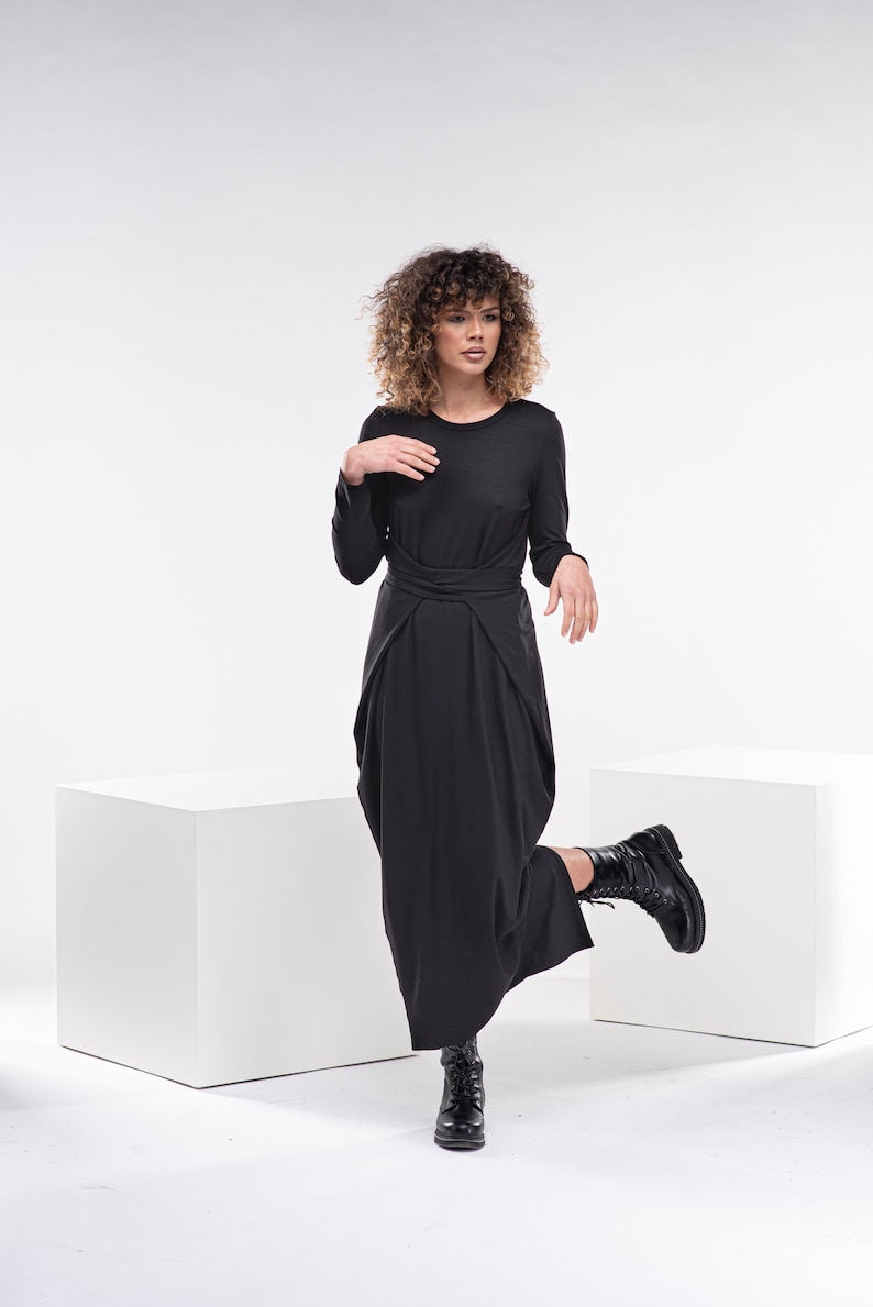May include: A woman wearing a black long-sleeved dress with a knotted waist. She is standing on one leg with her other leg bent at the knee and her foot resting on a white cube. The background is white.