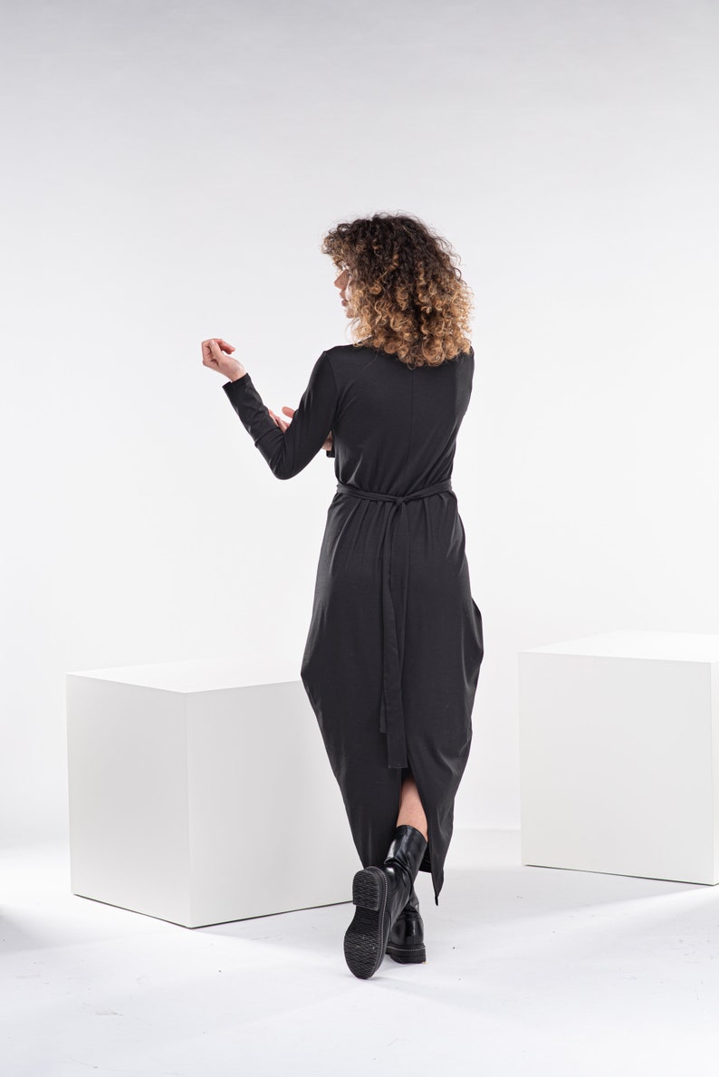 May include: A black long-sleeved dress with a tie at the waist. The dress has a high neckline and a long, flowing skirt. The model is wearing black boots.