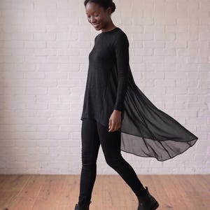 Op de afbeelding: Een zwarte tuniek met lange mouwen, een voorzijde van effen breiwerk en een transparant, vloeiend achterpand van chiffon dat uitloopt in een asymmetrische zoom, gecombineerd met een nauwsluitende zwarte broek en zwarte leren enkellaarsjes.
