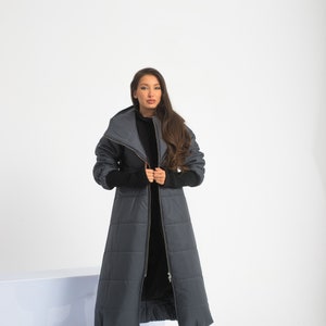 May include: A long, dark gray puffer coat with a high collar and a zipper closure. The coat has a quilted design and is worn by a person with long brown hair.