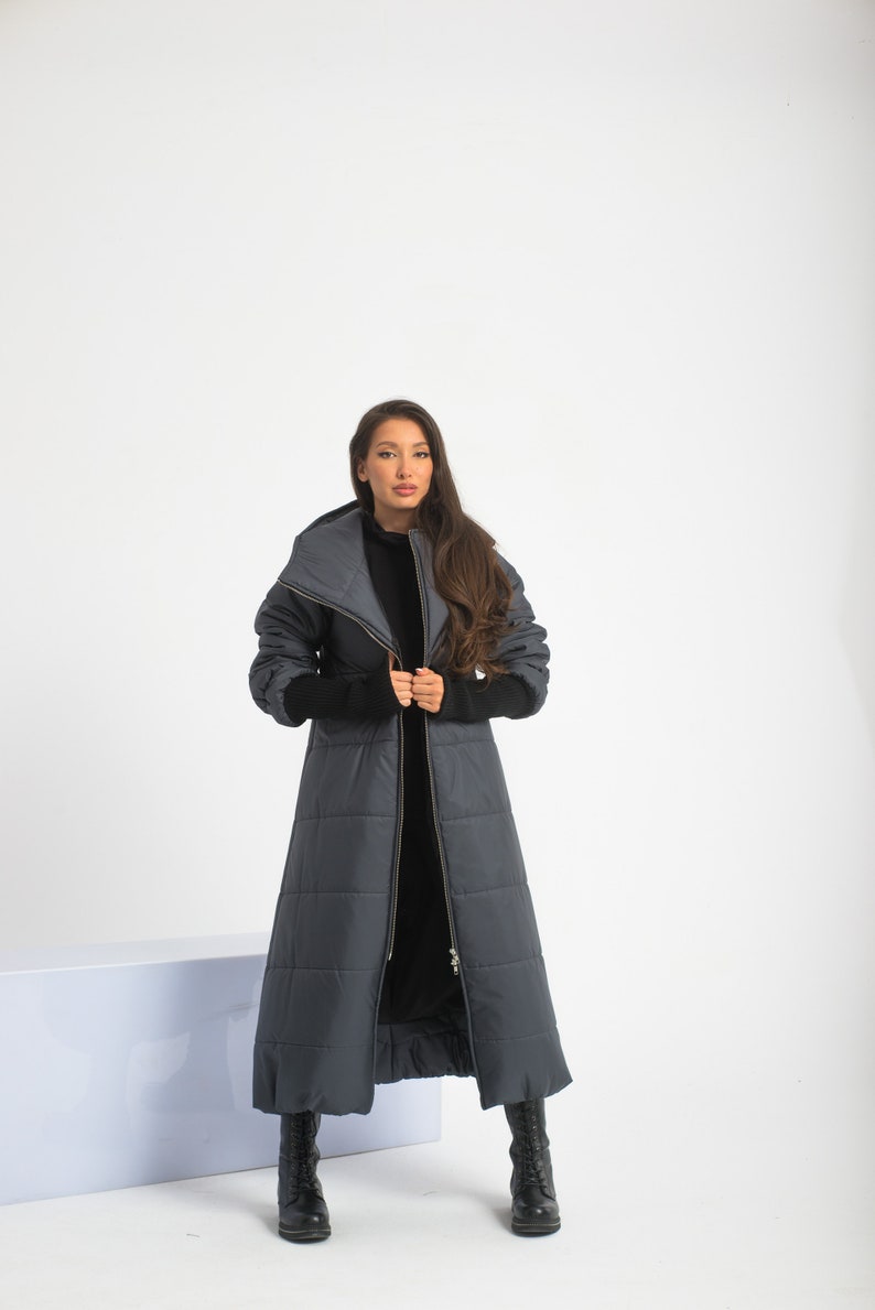 May include: A long, dark gray puffer coat with a high collar and a zipper closure. The coat has a quilted design and is worn by a person with long brown hair.