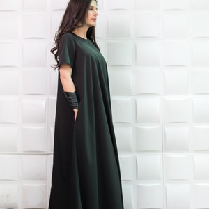 K&ouml;nnte beinhalten: Eine Frau tr&auml;gt ein langes, schwarzes, flie&szlig;endes Kleid mit kurzen &Auml;rmeln. Sie steht vor einer wei&szlig;en Wand mit einem strukturierten Muster.