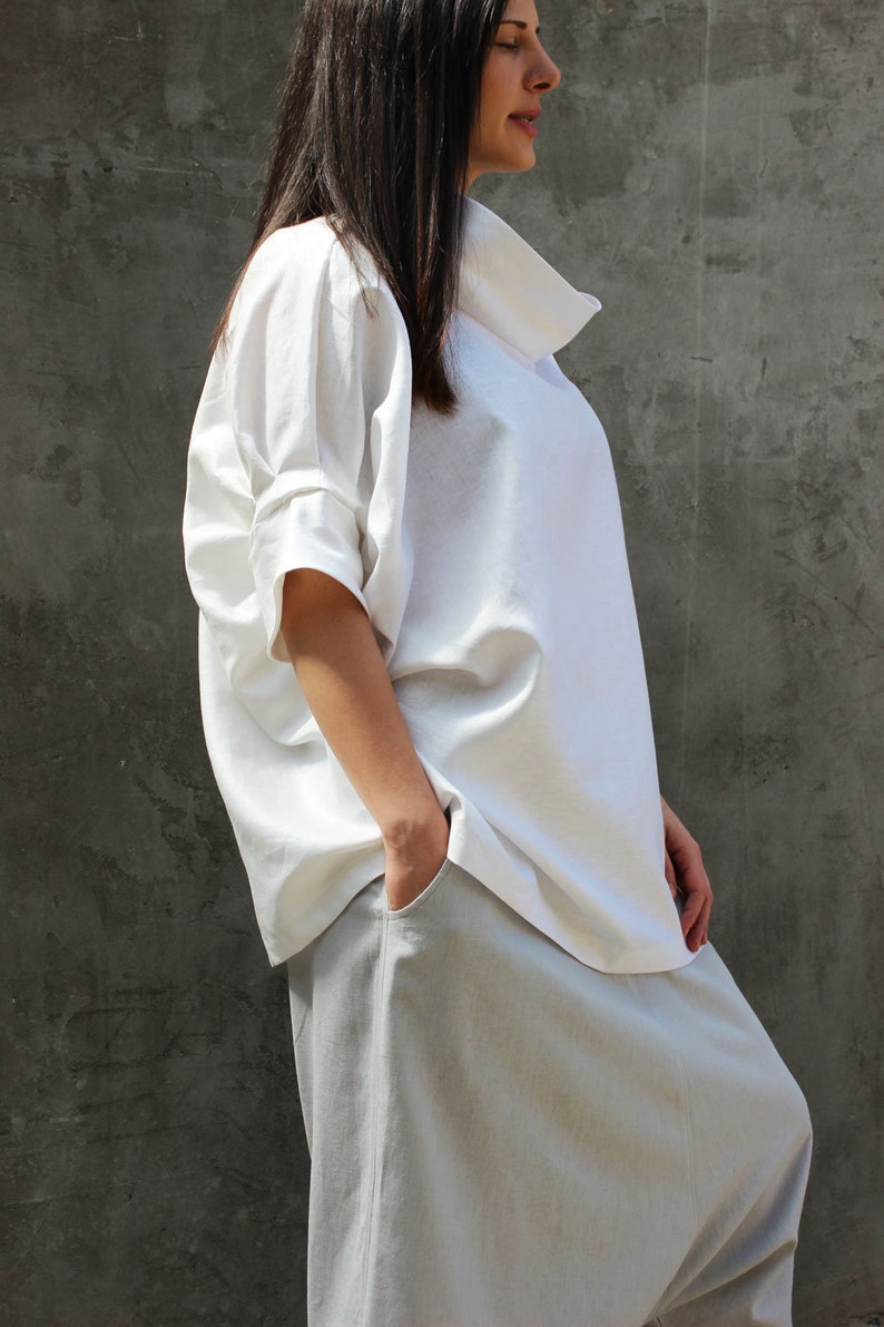 May include: A white linen shirt with a relaxed fit and a high, folded collar. The shirt is paired with light gray linen pants with a wide leg and a loose fit.