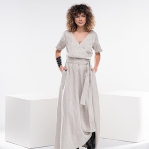 May include: A woman wearing a light beige linen wrap dress with short sleeves and a self-tie belt. She is standing on a white background.