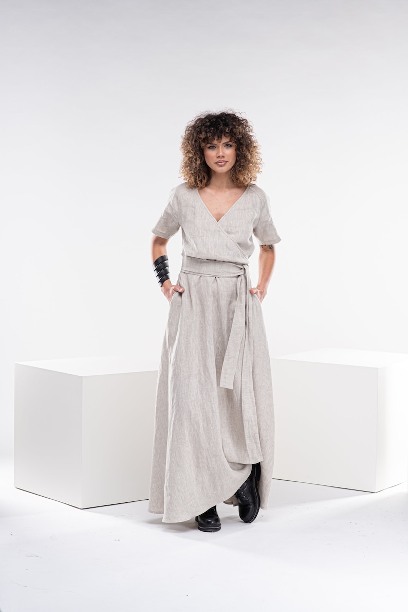 May include: A woman wearing a light beige linen wrap dress with short sleeves and a self-tie belt. She is standing on a white background.