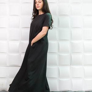 K&ouml;nnte beinhalten: Eine Frau tr&auml;gt ein schwarzes Maxikleid mit kurzen &Auml;rmeln und Taschen. Das Kleid ist flie&szlig;end und hat eine lockere Passform. Sie steht vor einer wei&szlig;en Wand mit einem strukturierten Muster.
