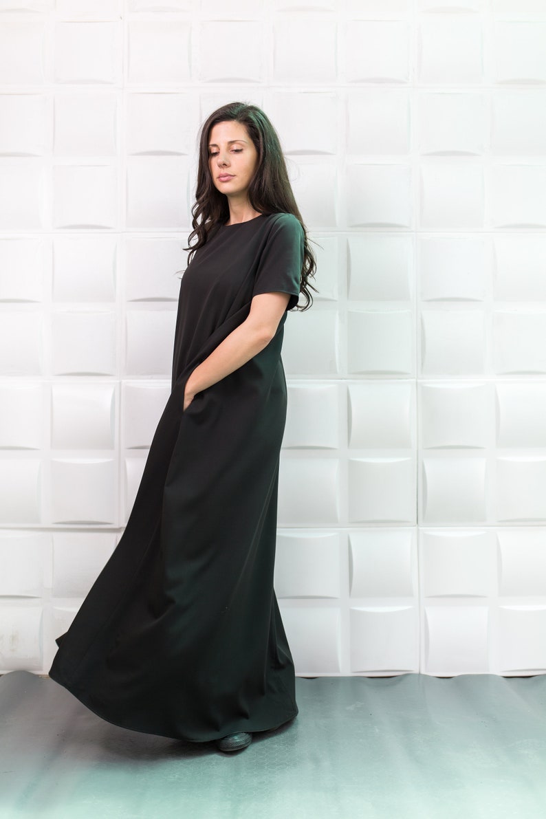 K&ouml;nnte beinhalten: Eine Frau tr&auml;gt ein schwarzes Maxikleid mit kurzen &Auml;rmeln und Taschen. Das Kleid ist flie&szlig;end und hat eine lockere Passform. Sie steht vor einer wei&szlig;en Wand mit einem strukturierten Muster.