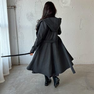 Puede incluir: Una larga gabardina gris oscura con capucha y cintur&oacute;n. La gabardina tiene una silueta fluida y acampanada.