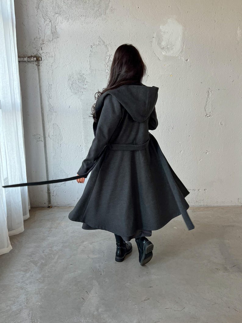 Puede incluir: Una larga gabardina gris oscura con capucha y cintur&oacute;n. La gabardina tiene una silueta fluida y acampanada.