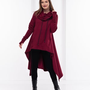 May include: A burgundy hooded sweatshirt with a long, asymmetrical hemline. The sweatshirt has a drawstring hood and long sleeves with thumbholes. The model is wearing black leggings and black boots.
