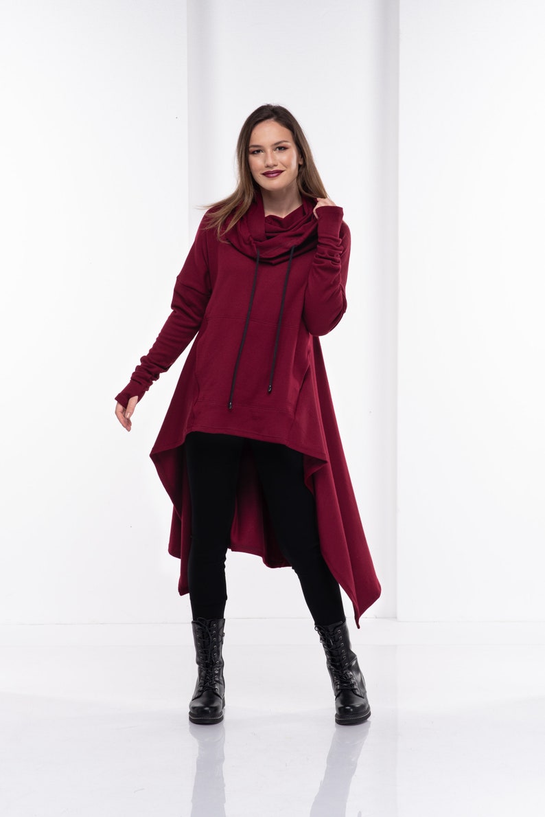 May include: A burgundy hooded sweatshirt with a long, asymmetrical hemline. The sweatshirt has a drawstring hood and long sleeves with thumbholes. The model is wearing black leggings and black boots.