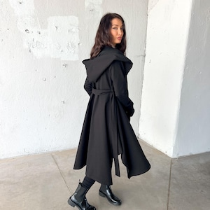 May include: A long, black hooded coat with a flowing skirt and a matching belt. The coat is worn with black boots and leggings. The coat's design features a dramatic, oversized hood and a wrap-around belt at the waist.