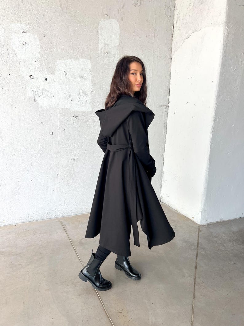 May include: A long, black hooded coat with a flowing skirt and a matching belt. The coat is worn with black boots and leggings. The coat's design features a dramatic, oversized hood and a wrap-around belt at the waist.