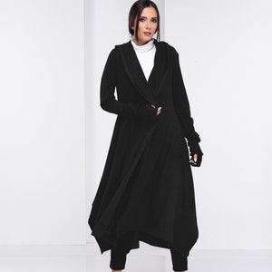 May include: A black long-sleeved hooded cardigan with a draped asymmetrical hem. The cardigan is worn over a white turtleneck shirt and black pants. The model is wearing silver shoes.