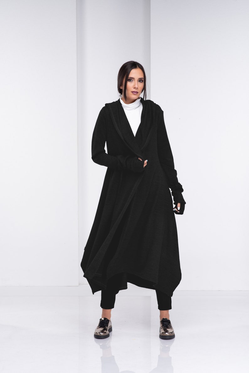 May include: A black long-sleeved hooded cardigan with a draped asymmetrical hem. The cardigan is worn over a white turtleneck shirt and black pants. The model is wearing silver shoes.