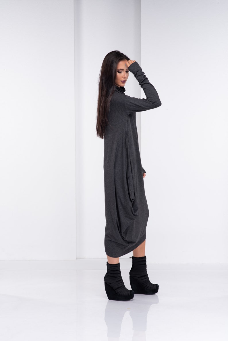 May include: A charcoal gray long-sleeved dress with a loose, flowing fit. The dress has a high neckline and a long, flowing skirt. The model is wearing black ankle boots.