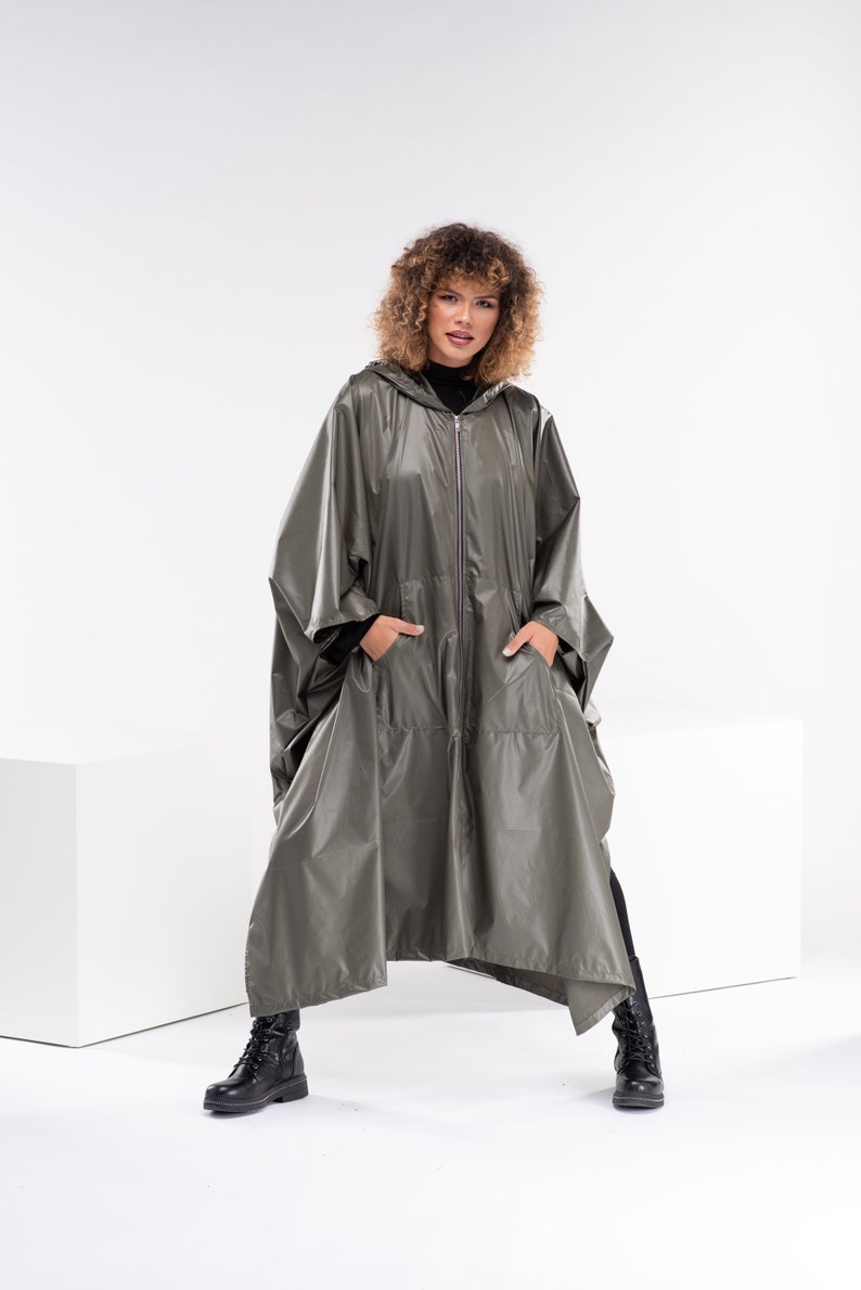 May include: A woman wearing a long, olive green hooded raincoat with a zipper closure. The raincoat has two large pockets and a loose, flowing fit. She is wearing black boots and has curly brown hair.